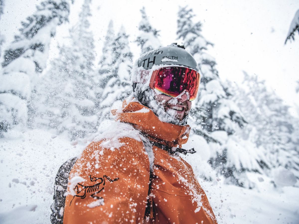A snowboarder in goggles and helmet plunges through heavy snow in a mountain forest, spraying white flakes.
