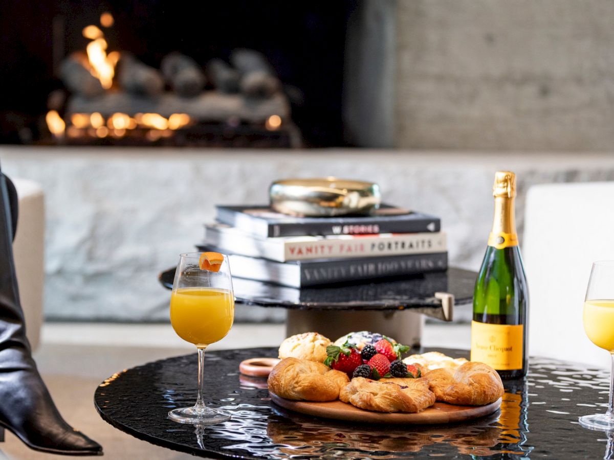 Brunch on a table with orange juice in glasses, a champagne bottle, pastries and fruit, with books and candles in the background.