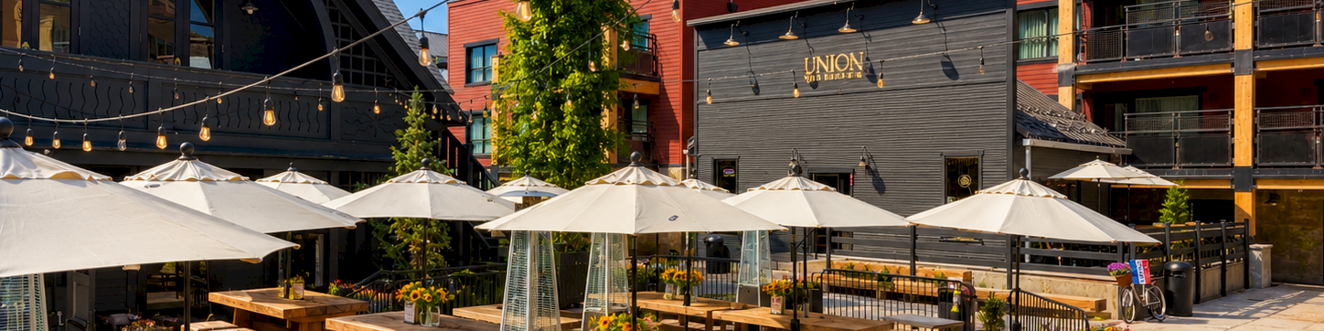 A sunny outdoor dining area with wooden benches, tables, and white umbrellas, surrounded by modern brick buildings and string lights.