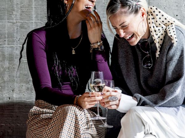 Two women are laughing and toasting with wine glasses while sitting together against a gray wall.