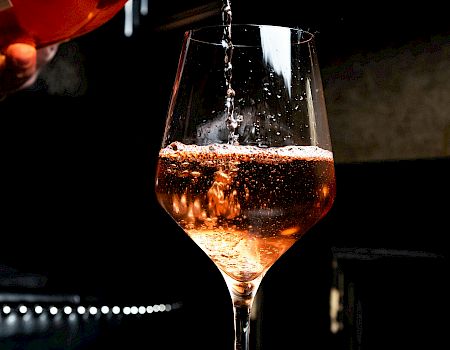 A bartender pours a dark amber drink from a bottle into a stemmed glass, sparkling as it fills.