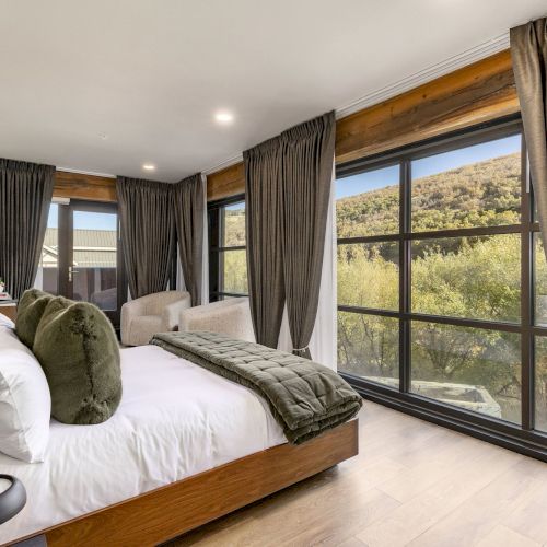 A cozy bedroom with a white bed, plush green pillows, floor-to-ceiling gray curtains, and large windows overlooking hills, serene.