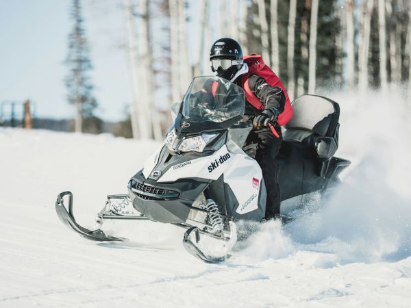 A rider in helmet charges through snowy forest on a Ski-Doo snowmobile, kicking up snow spray behind it.