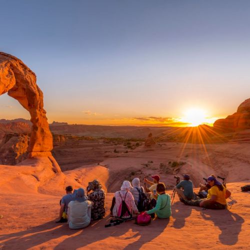 People sit and watch a desert sunset at a natural rock arch, with warm sunrays cutting through the canyon.