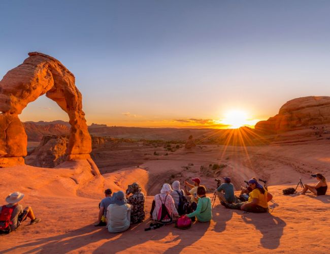 People sit and watch a desert sunset at a natural rock arch, with warm sunrays cutting through the canyon.