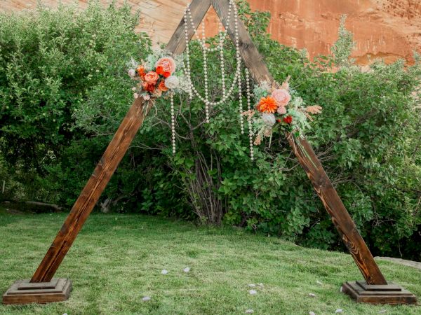 An outdoor ceremony arch of wood and pearls, decorated with flowers on a grassy lawn, backed by a red rock cliff.