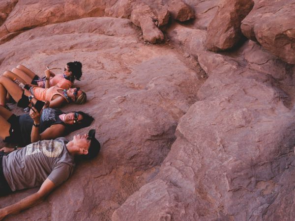 A group of friends lies on reddish rocks, chatting and using phones, soaking up the sun and scenic desert surroundings.